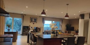 Un vaste espace de vie comprenant un grand îlot central, une table à manger et un coin salon. De nombreuses fenêtres laissent entrer la lumière naturelle et offrent une vue sur l’extérieur.