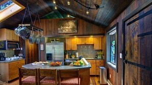 A rustic kitchen with wooden cabinets, a central island with chairs, hanging pots, and a refrigerator.