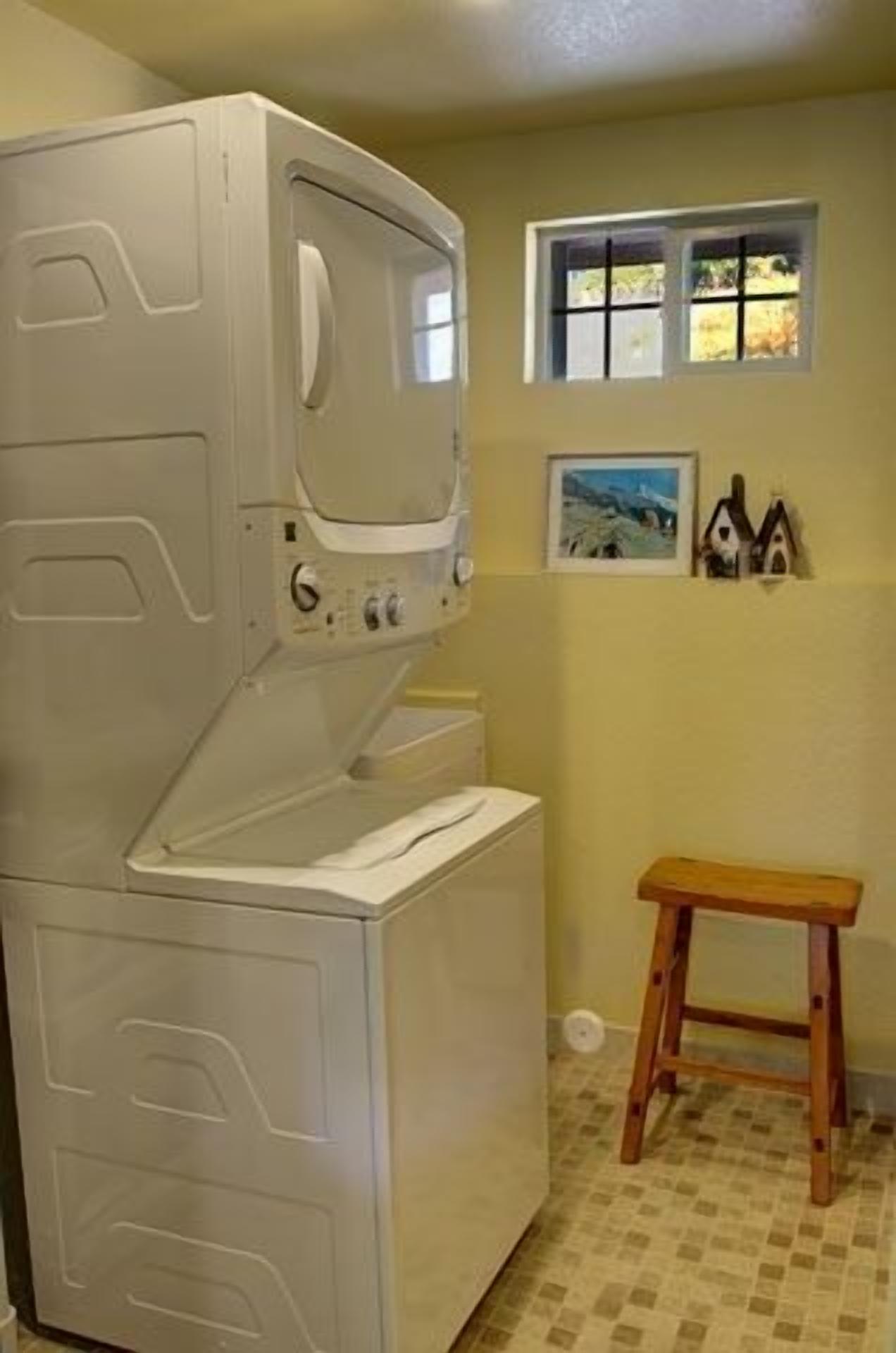 A laundry room with stacked washer and dryer, a wooden stool, and a window with a view of trees.