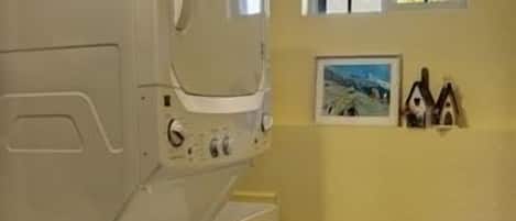A laundry room with stacked washer and dryer, a wooden stool, and a window with a view of trees.