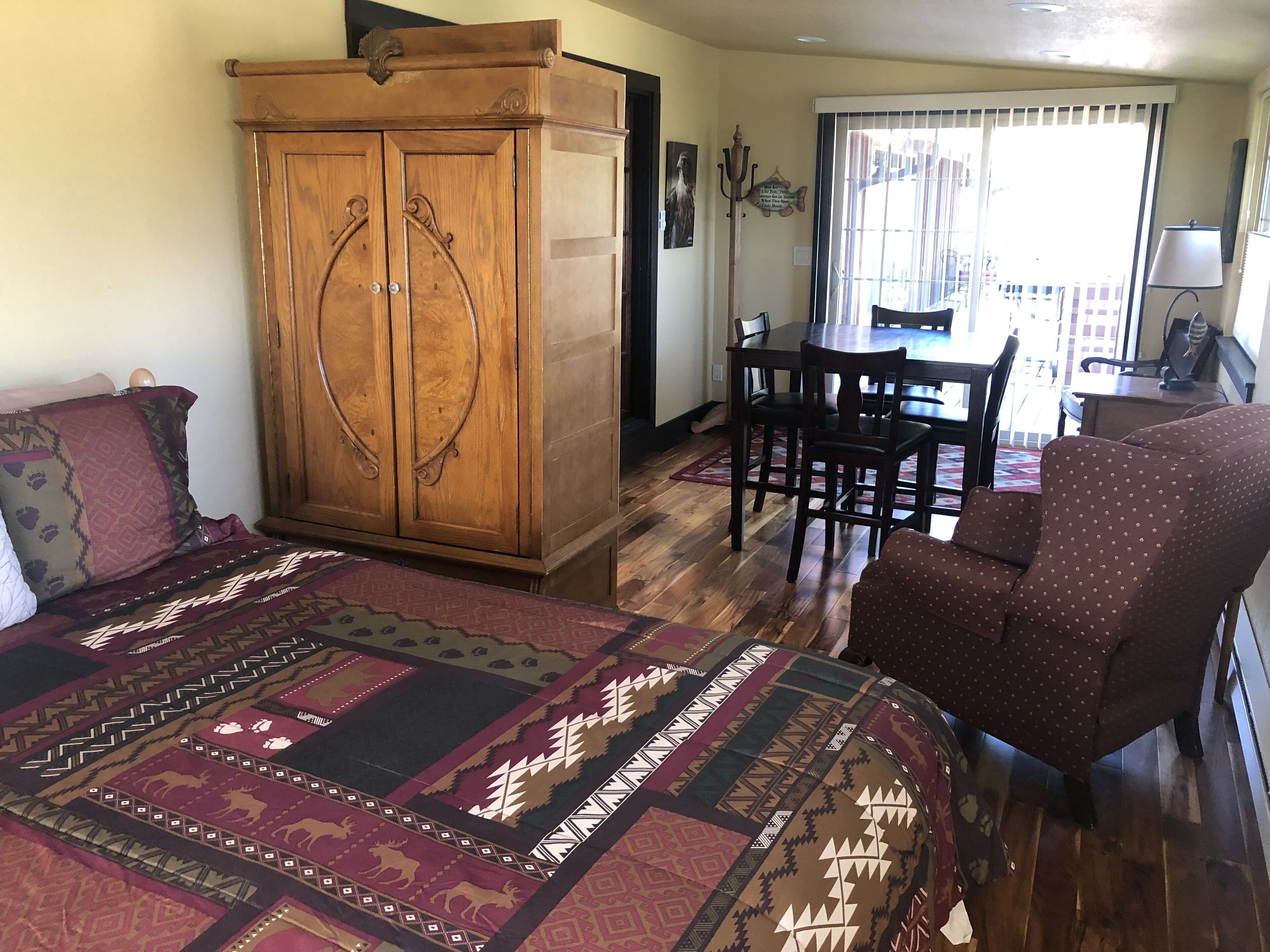 A room with a large wooden wardrobe, a patterned rug, a dining table with chairs, and a polka-dotted armchair.