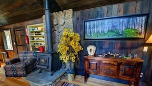A cozy living room with a stone fireplace, a plaid sofa, a wooden cabinet, and a framed forest painting.