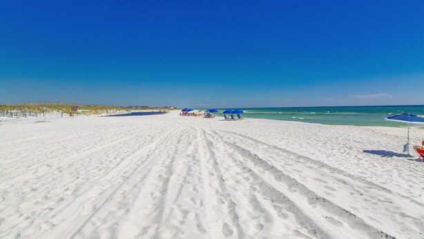 A sandy beach with footprints leading to the water, a few umbrellas, and a clear blue sky.