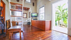 A spacious room with wooden flooring, a large window with a view of greenery, a television, and a wooden bench. - Historic Old Town  Key West Cigarmaker's Cottage - Cigar House, Sleeps 4 (Key West)