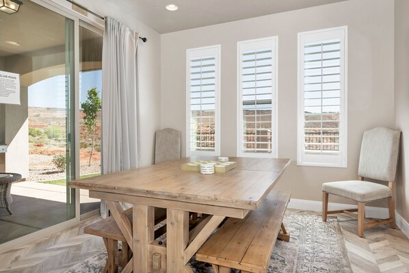 Un espace repas avec une table et des bancs en bois, de grandes fenêtres équipées de stores et une vue sur un paysage désertique.