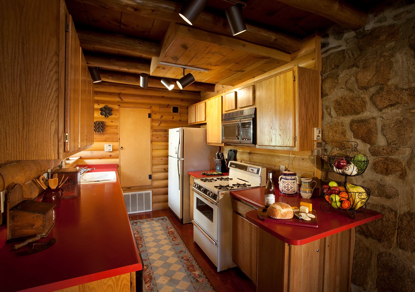 Una cocina rústica con armarios de madera, encimera roja, estufa, refrigerador y pared de piedra.