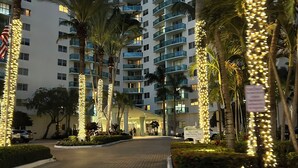 Een meerlaags hotel met een modern ontwerp, voorzien van balkons op elke verdieping, een verhard entreegebied en palmbomen.