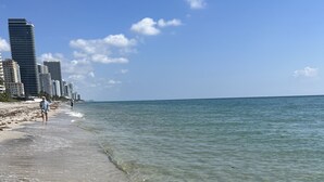 Een strand met helder ondiep water, een zandstrand en hoge gebouwen op de achtergrond.