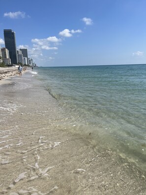 A beach with clear shallow waters, a sandy shore, and high-rise buildings in the background.