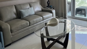Een moderne woonkamer met een leren bank, een glazen salontafel en een groot raam met gordijnen.