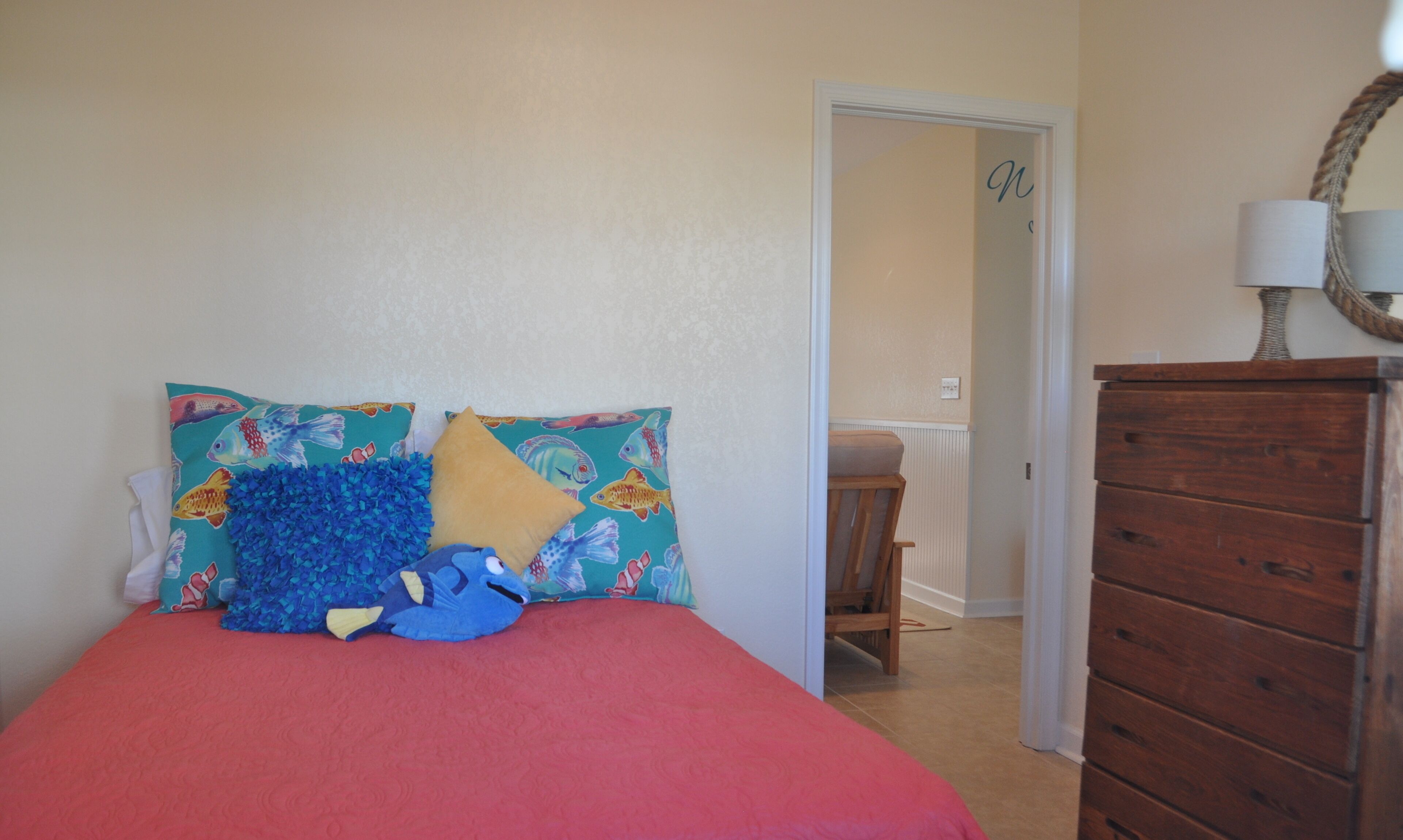 A bedroom with a bed, colorful pillows, a wooden dresser, and a lamp.