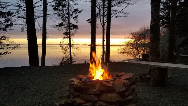 Una fogata en un hogar de piedra rodeada de un bosque, con un lago y un atardecer al fondo.