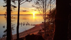 Un atardecer sobre un lago tranquilo con una playa arenosa y árboles en primer plano.