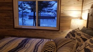 Una habitación de cabaña de madera con un ventanal grande que da a un lago tranquilo y árboles, una cama con ropa de cama estampada y una lámpara de noche.