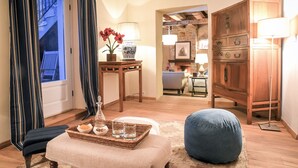 Una sala de estar acogedora con un sofá, un reposapiés y una mesa con una bandeja de aperitivos y bebidas. Hay un aparador de madera y una lámpara sobre una mesita auxiliar.