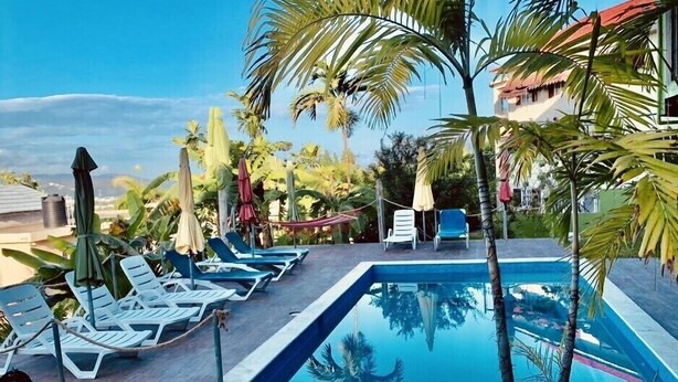 A swimming pool with lounge chairs and umbrellas, surrounded by palm trees and a building with a red roof. - GD Rooftop Lounge ocean view, salt water pool with mountain view, 1bdrm (West Gate Hills, Montego Bay)