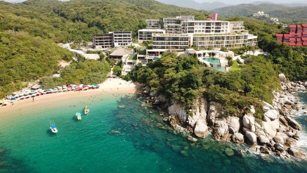 Vista aérea de un complejo turístico con varias edificaciones, una playa y aguas turquesas cristalinas.