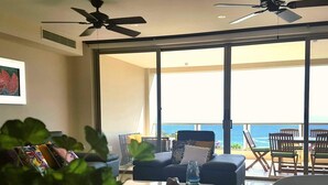 Un salón moderno con un amplio ventanal corredera que da a un balcón con vista al océano. Hay ventiladores de techo, una mesa de centro con un sombrero y un vaso, y una estantería con libros.