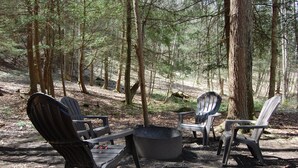 A campsite with Adirondack chairs arranged around a fire pit in a forested area. - Dreamcatcher-Harvest Moon Cottages (Rockbridge)