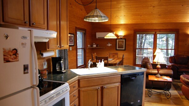 A kitchen with wooden cabinets, a refrigerator, a stove, and a dishwasher. There is a dining table with chairs and a couch in the living area. - Dreamcatcher-Harvest Moon Cottages (Rockbridge)