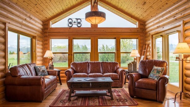 A log cabin interior with leather sofas, a central coffee table, and large windows offering a view of the outdoors. - Gorgeous Saddlehorn Cabin - Amazing Teton Views (Driggs)