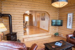 A log cabin interior with a wooden floor, leather furniture, a television mounted on the wall, and a fireplace.