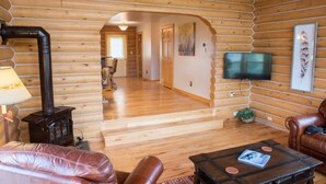 A log cabin interior with a wooden floor, leather furniture, a TV mounted on the wall, and a fireplace. - Gorgeous Saddlehorn Cabin - Amazing Teton Views (Driggs)
