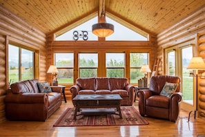 Interior de una cabaña de madera con techo alto, muebles de cuero y grandes ventanales con vistas al exterior.