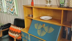 A room with a wooden cabinet with blue and gold decorative panels, a black leather chair, a patterned throw pillow, and a window with geometric curtains. - Kims Cozy Rustic Cabin 5 min. from Cody (Cody)