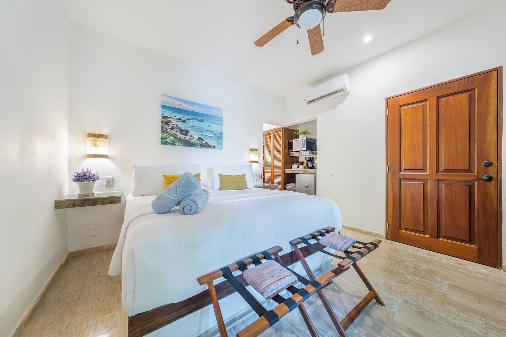 A bedroom with a bed, a wooden wardrobe, a ceiling fan, a wall-mounted air conditioner, and a painting of a beach scene.
