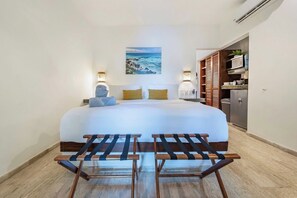 A hotel room with a bed, two bedside lamps, a painting of a beach, a small table with a towel, and a kitchenette.