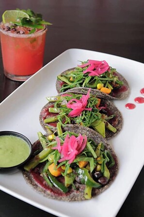 A plate with three tacos topped with vegetables and a green sauce, a glass of red drink, and a small bowl of green sauce.