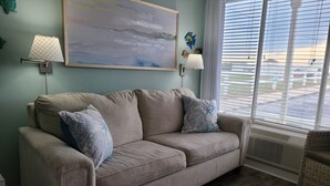 A beige sofa with patterned pillows, a wall-mounted painting, and a window with blinds. - BEACH TIME!  Ocean side! Private area, no crowds - Steps from sand.  Downstairs. (Atlantic Beach)