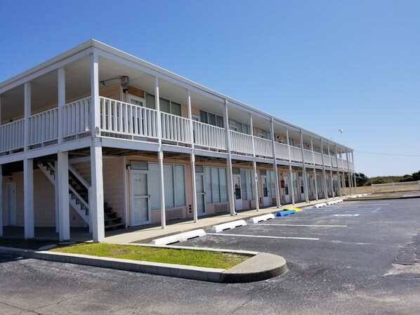 A two-story motel with a balcony on the upper floor, a parking lot in front, and a clear blue sky.
