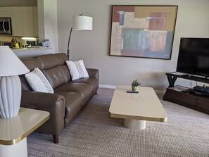 A modern living room with a brown leather sofa, a white coffee table, a lamp, a flat-screen TV, a painting, and a small plant.