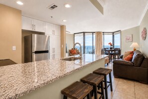 A modern kitchen with a granite countertop, stainless steel appliances, and a dining area with a view. - Renovated 8th-Floor Beachfront Condo with Stunning Sunset Views (Panama City Beach)