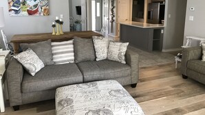 A living room with a grey sofa, ottoman, and a dining table with chairs. There is a kitchen area with a refrigerator and a hanging light fixture. - 5 Bedroom/3 Bath Emerald Coast Beach Cottage in gated resort community (Destin)