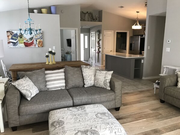 A living room with a grey sofa, ottoman, and a dining table with chairs. There is a kitchen area with a refrigerator and a hanging light fixture. - 5 Bedroom/3 Bath Emerald Coast Beach Cottage in gated resort community (Destin)