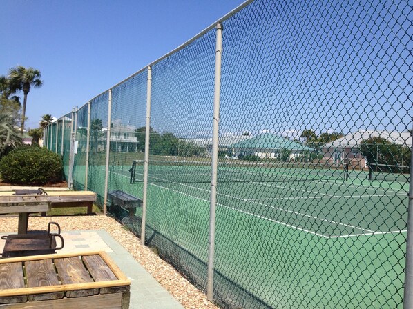 Una cancha de tenis rodeada por una cerca de malla metálica, con un banco de madera y una mesa de picnic cerca.
