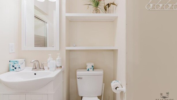 A bathroom with a white sink, a toilet, and a mirror above the sink. There are shelves above the toilet with decorative items.