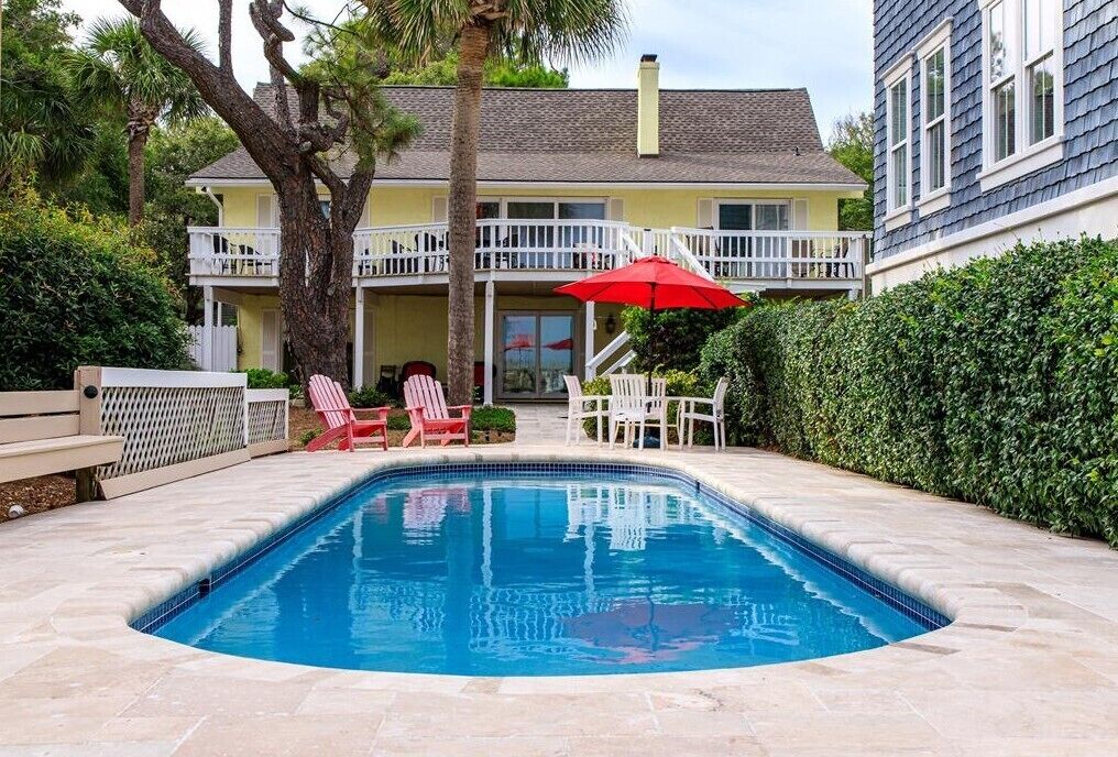 Una piscina con una sombrilla roja, sillas de deck blancas y un edificio de dos pisos con balcones.