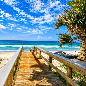 Ein hölzerner Steg führt zu einem Strand mit klarem, blauem Wasser und einer Palme.