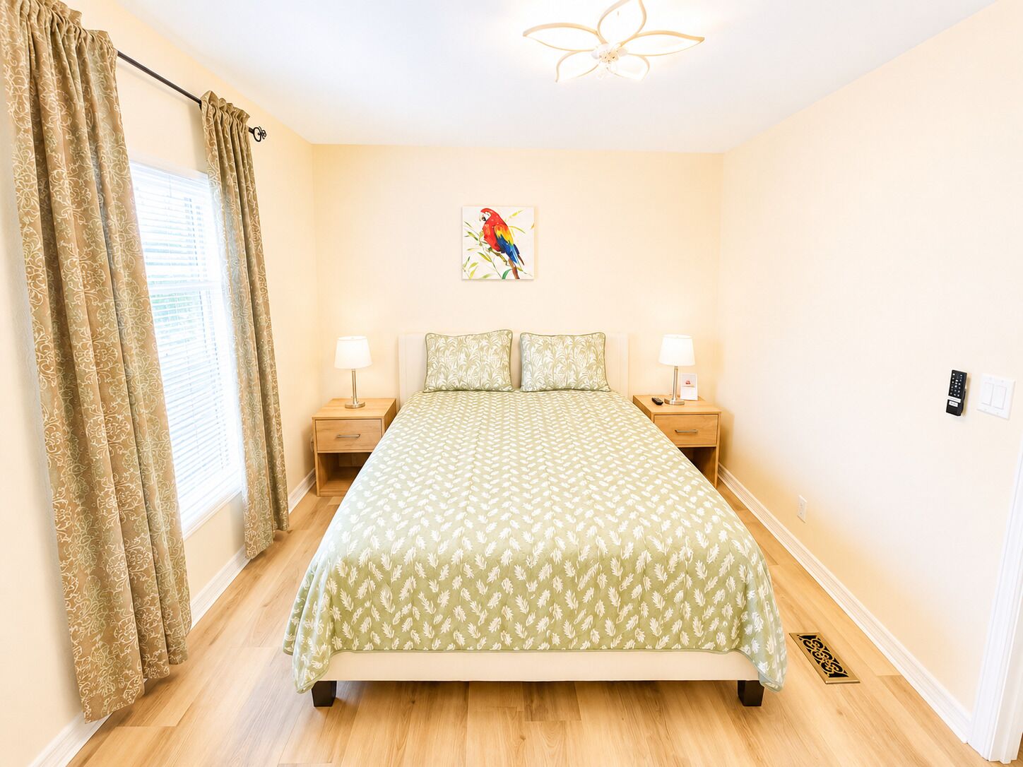 A bedroom with a bed, two bedside tables, a ceiling fan, a window with curtains, and a wall-mounted artwork.