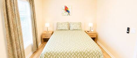 A bedroom with a bed, two bedside tables, a ceiling fan, a window with curtains, and a wall-mounted artwork.