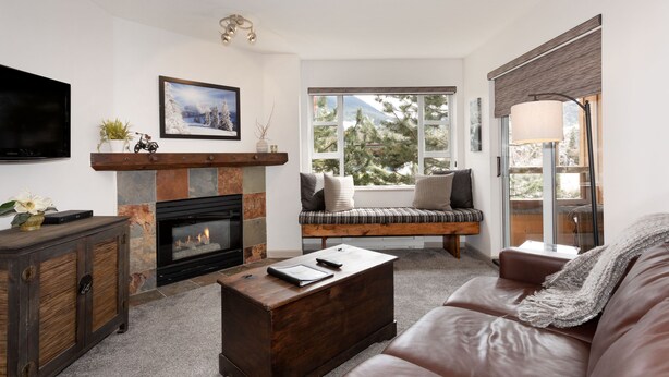 A living room with a fireplace, a sofa, a coffee table, and a window with a view of trees. - Mountain Views in Whistler Village (Whistler)