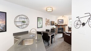 A modern kitchen with a central island, dining area with chairs, a fireplace, and a bicycle mounted on the wall. - Mountain Views in Whistler Village (Whistler)
