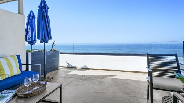 A balcony with a view of the sea, a blue sofa, two blue umbrellas, and a wooden chair.