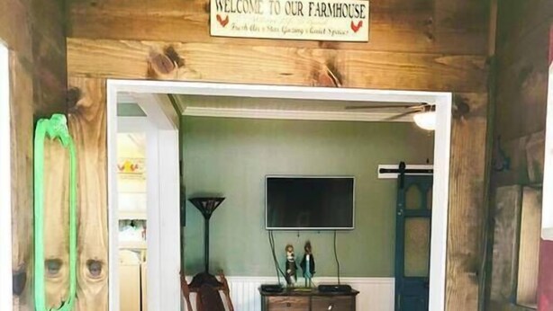 Ein Holzinterieur mit einem Schild 'Welcome to our Farmhouse', einem Holzstuhl, einer Kommode und einem flachbildfernseher an der Wand.