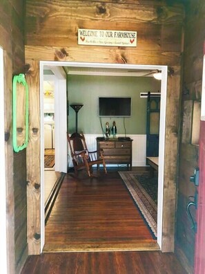 Un interior de madera con un letrero que dice 'Welcome to our Farmhouse', una silla de madera, un aparador y un televisor de pantalla plana montado en la pared.
