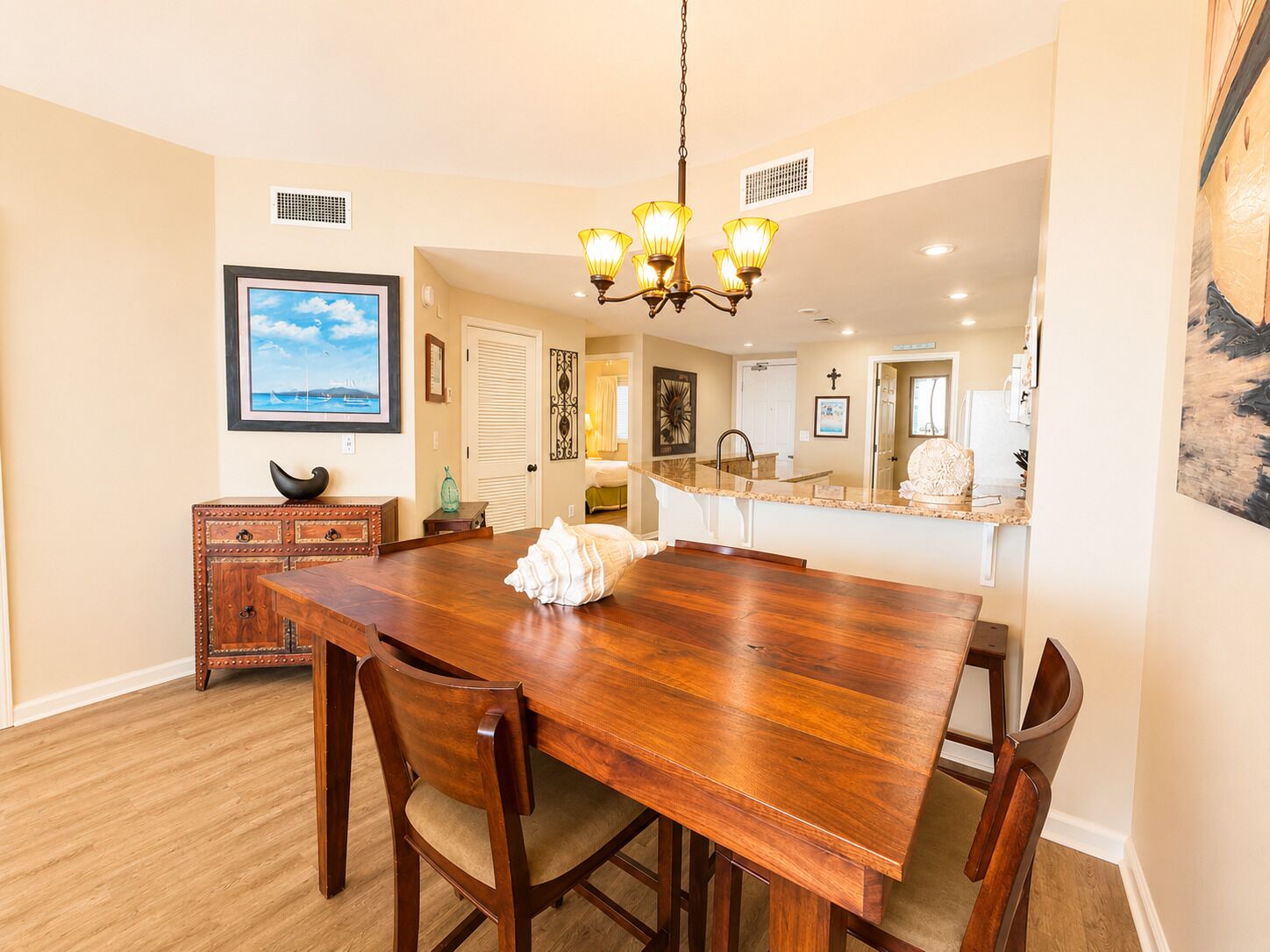 A dining area with a wooden table, chairs, a chandelier, and a view of the kitchen.